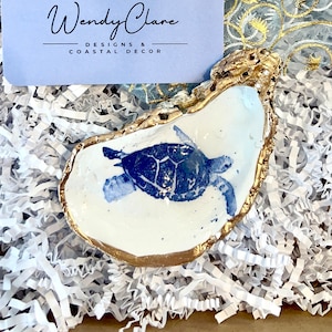 May include: A decorative oyster shell painted with a blue sea turtle, featuring a gold-painted edge. A light blue business card displays the text Wendy Clare DESIGNS & COASTAL DECOR. The items rest on white paper shred inside a cardboard box.