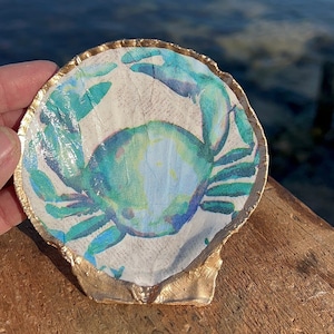 May include: A white seashell with a gold rim, painted with a blue and green crab. The seashell is sitting on a wooden surface.