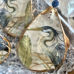 May include: Two oyster shell ornaments with gold trim, each featuring a blue heron bird illustration and a green leafy background. The shells are displayed on a white and gray crinkled tissue paper.