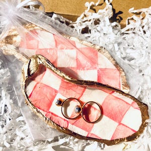 May include: Two gold rings and a pair of stud earrings rest on a pink and white checkered oyster shell. The shell has a gold gilded edge. A second shell and a sheer bag are visible in a box with white paper filler.