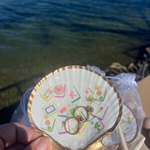 May include: A white seashell ring dish with a gold-painted rim and base, decorated with colorful, patterned tiles. Two gold rings with gemstones rest inside the dish. A starfish and a small bag are also visible.