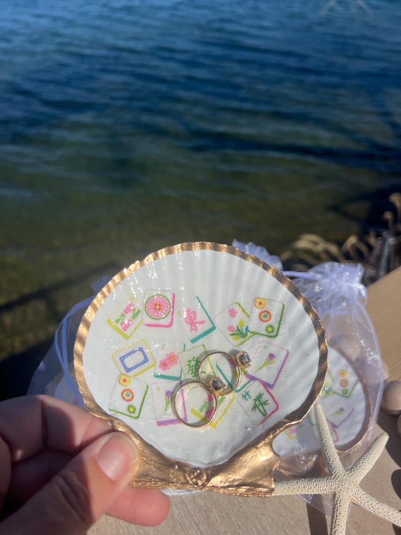 May include: A white seashell ring dish with a gold-painted rim and base, decorated with colorful, patterned tiles. Two gold rings with gemstones rest inside the dish. A starfish and a small bag are also visible.