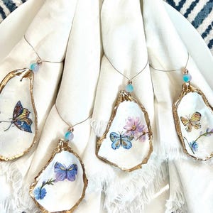 K&ouml;nnte beinhalten: Vier wei&szlig;e Austernmuscheln mit goldener Verzierung, jede mit einem anderen bemalten Schmetterlingsdesign. Die Muscheln sind mit einem d&uuml;nnen Draht und einer kleinen blauen Perle an wei&szlig;en Servietten befestigt.