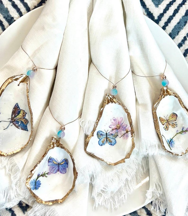 K&ouml;nnte beinhalten: Vier wei&szlig;e Austernmuscheln mit goldener Verzierung, jede mit einem anderen bemalten Schmetterlingsdesign. Die Muscheln sind mit einem d&uuml;nnen Draht und einer kleinen blauen Perle an wei&szlig;en Servietten befestigt.