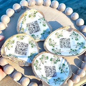 May include: Four decorative oyster shells with printed floral designs and QR codes, arranged on a wooden tray with a beaded border. The shells have a gold-colored edge and are set against a blurred water background.