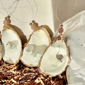 May include: Three gold-painted oyster shell ornaments with a white interior. Each ornament features a different animal illustration: a rhinoceros, an ostrich, and a warthog. The ornaments are strung with gold wire and have a gold bead at the top.