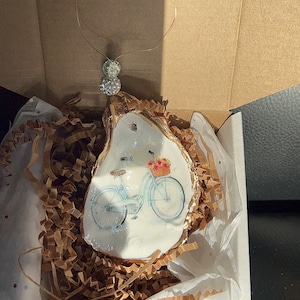 May include: A white oyster shell ornament with a gold rim, featuring a painted image of a blue bicycle with a basket of flowers. The ornament is packaged in a white box with brown paper shred.