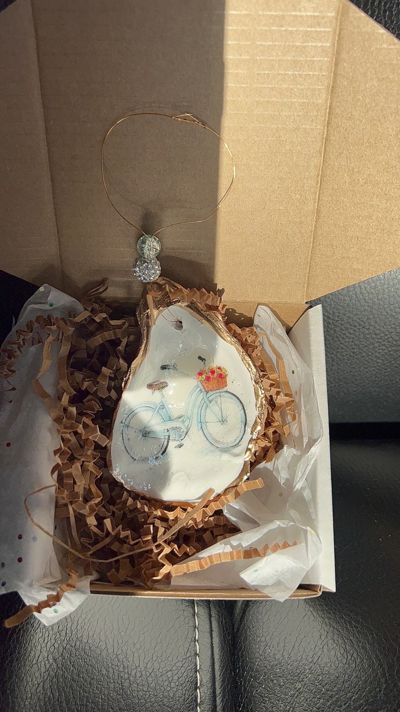 May include: A white oyster shell ornament with a gold rim, featuring a painted image of a blue bicycle with a basket of flowers. The ornament is packaged in a white box with brown paper shred.