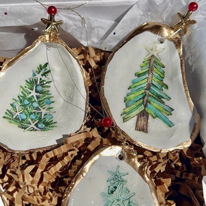 May include: Three oyster shell Christmas ornaments with gold trim. Each ornament features a different design of a Christmas tree: one with blue and green pine needles, one with green and white stripes, and one with a design of starfish.