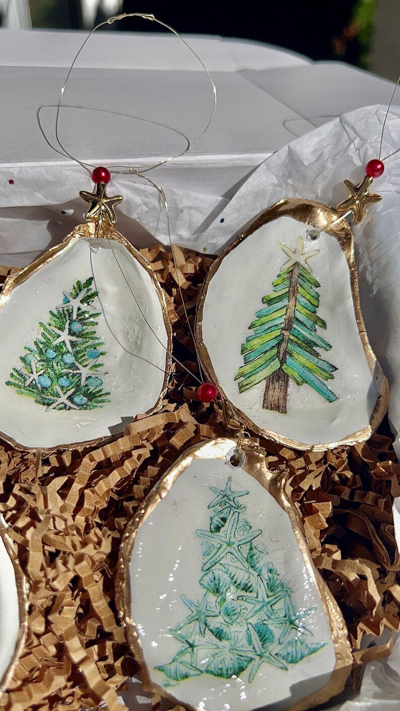 May include: Three oyster shell Christmas ornaments with gold trim. Each ornament features a different design of a Christmas tree: one with blue and green pine needles, one with green and white stripes, and one with a design of starfish.