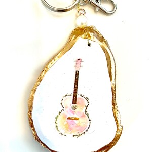 May include: A gold-painted oyster shell keychain with a painted image of a pink and orange guitar. The keychain has a silver ring and clasp.