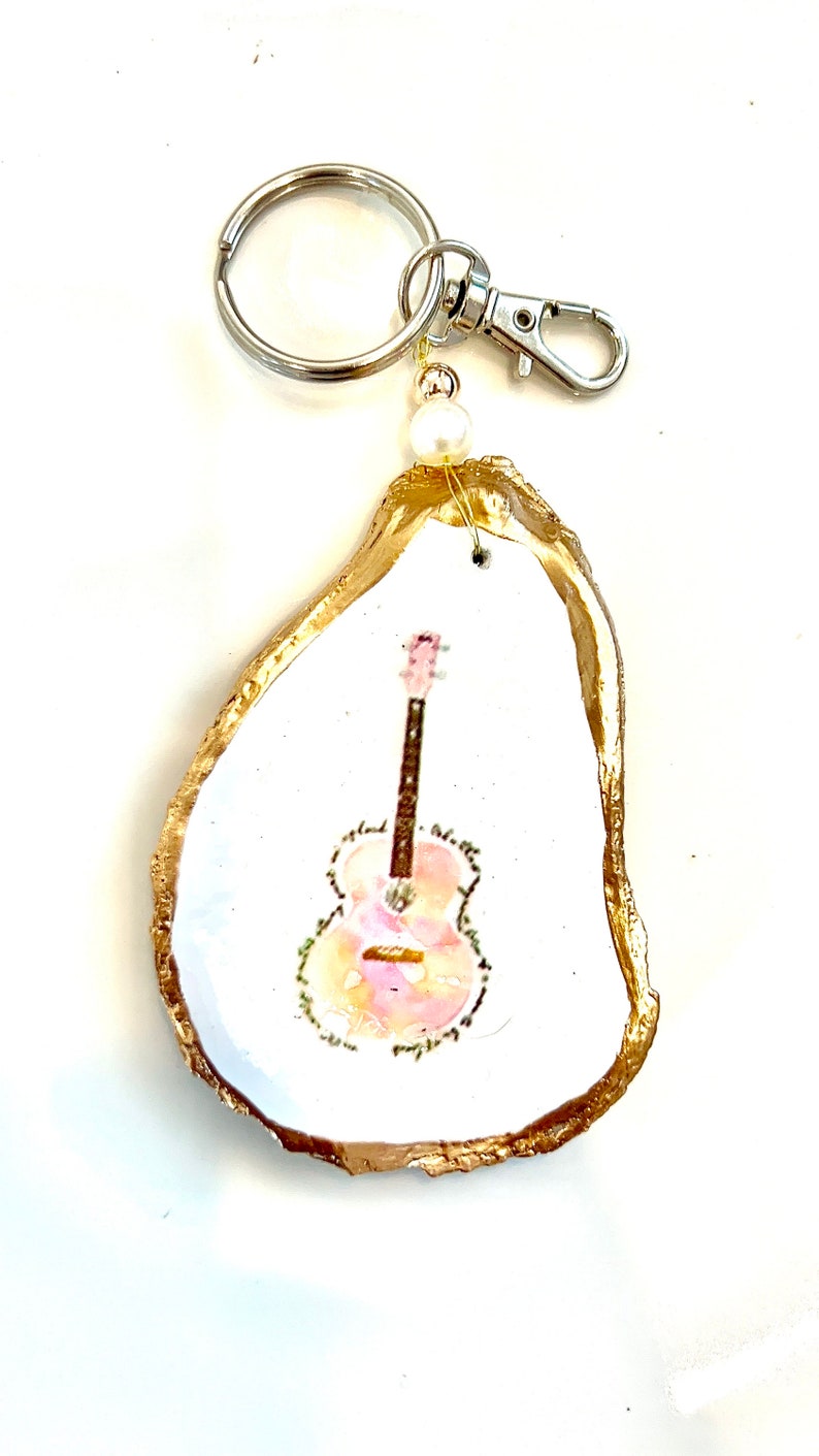 May include: A gold-painted oyster shell keychain with a painted image of a pink and orange guitar. The keychain has a silver ring and clasp.