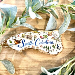 Op de afbeelding: Een wit keramiek ornament in de vorm van de staat South Carolina, versierd met kleurrijke illustraties van bezienswaardigheden en symbolen, waaronder een watermeloen, een golfclub, een vuurtoren en een palmettoboom. De tekst "South Carolina" is geschreven in blauwe cursieve letters.
