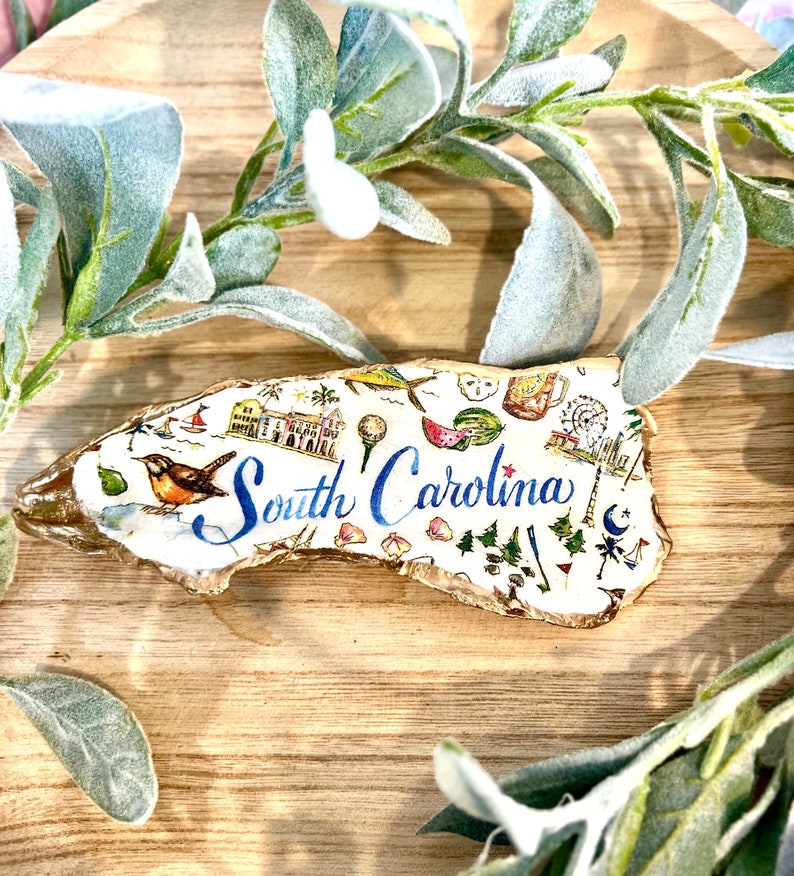 Op de afbeelding: Een wit keramiek ornament in de vorm van de staat South Carolina, versierd met kleurrijke illustraties van bezienswaardigheden en symbolen, waaronder een watermeloen, een golfclub, een vuurtoren en een palmettoboom. De tekst "South Carolina" is geschreven in blauwe cursieve letters.