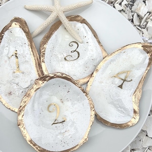 May include: A white plate holds four oyster shell dishes with gold rims and gold numerals 1, 2, 3, and A. A starfish rests on the plate. The shells are on a bed of white and gray shells.