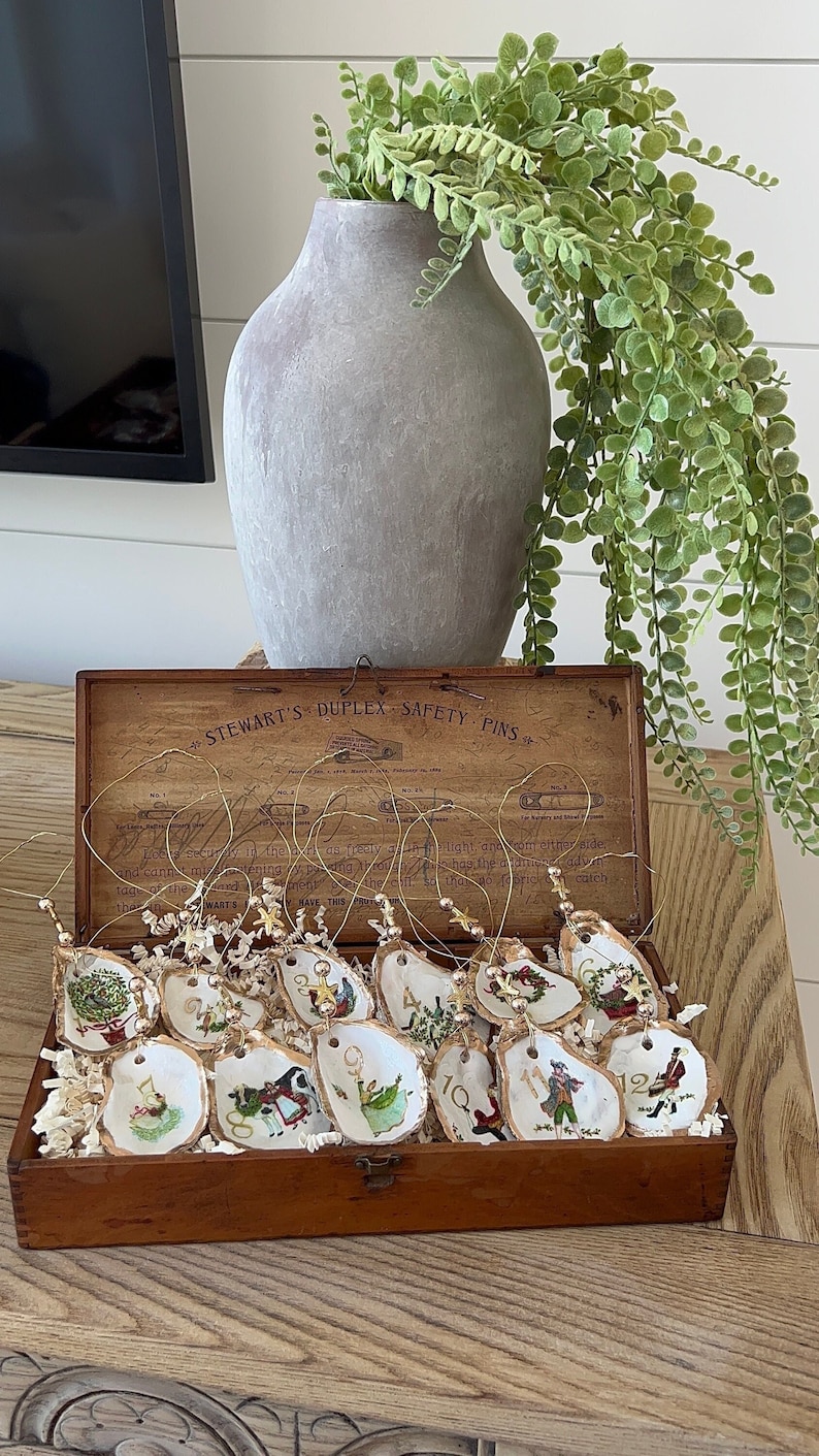 May include: A wooden box with a hinged lid, containing 12 oyster shell ornaments with gold wire hangers. Each ornament is painted with a different Christmas scene and numbered 1 through 12. The box is labeled "Stewart's Duplex Safety Pins".