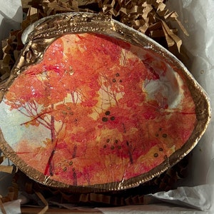 Puede incluir: Una concha de mar con un borde dorado y una escena de otoño pintada en su interior. La escena presenta árboles con hojas rojas, naranjas y amarillas, y un cielo azul claro.