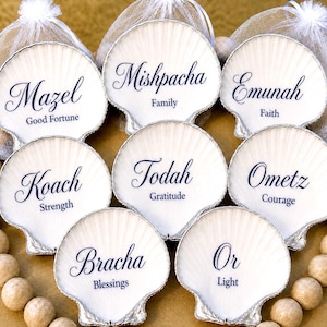 Peut inclure: Huit coquilles Saint-Jacques blanches aux bords peints en argent, chacune portant un mot hébreu en écriture bleue foncée avec sa traduction en anglais. Les mots incluent Mazel, Mishpacha, Emunah, Koach, Todah, Ometz, Bracha et Or.
