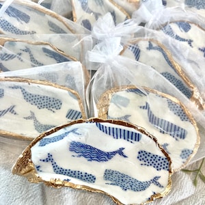 May include: A set of small, white ceramic oyster shells with a blue and white whale pattern. The shells are trimmed with gold leaf and are packaged in individual clear organza bags.