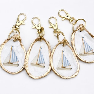 May include: Four keychains made from natural oyster shells, each featuring a hand-painted sailboat with blue and white striped sails. The edges of the shells are gilded in gold, and each is attached to a gold-toned metal lobster clasp.