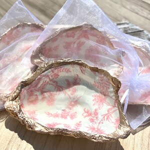 Può includere: Tre piatti a forma di conchiglia di ostrica con un motivo floreale rosa e bianco e un bordo dorato. I piatti sono esposti su un tessuto bianco trasparente.