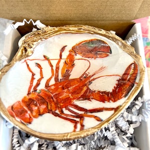 Op de afbeelding: Een witte keramische schaal met een gouden rand, met een handgeschilderde illustratie van een rode kreeft. De schaal wordt gepresenteerd op een bed van wit kreukelpapier.