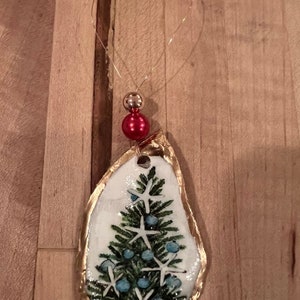 May include: A gold-trimmed oyster shell ornament with a painted Christmas tree and blue and white starfish. The ornament has a red bead at the top and a gold wire loop for hanging.