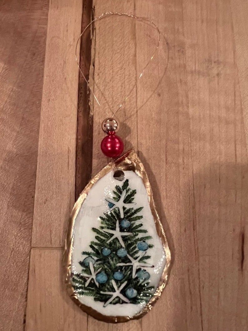 May include: A gold-trimmed oyster shell ornament with a painted Christmas tree and blue and white starfish. The ornament has a red bead at the top and a gold wire loop for hanging.