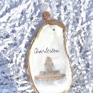Op de afbeelding: Een witte oester schelp ornament met gouden rand, met een geschilderd beeld van de fontein in Charleston, South Carolina. De ornament is versierd met het woord "Charleston" en een gouden touwtje.