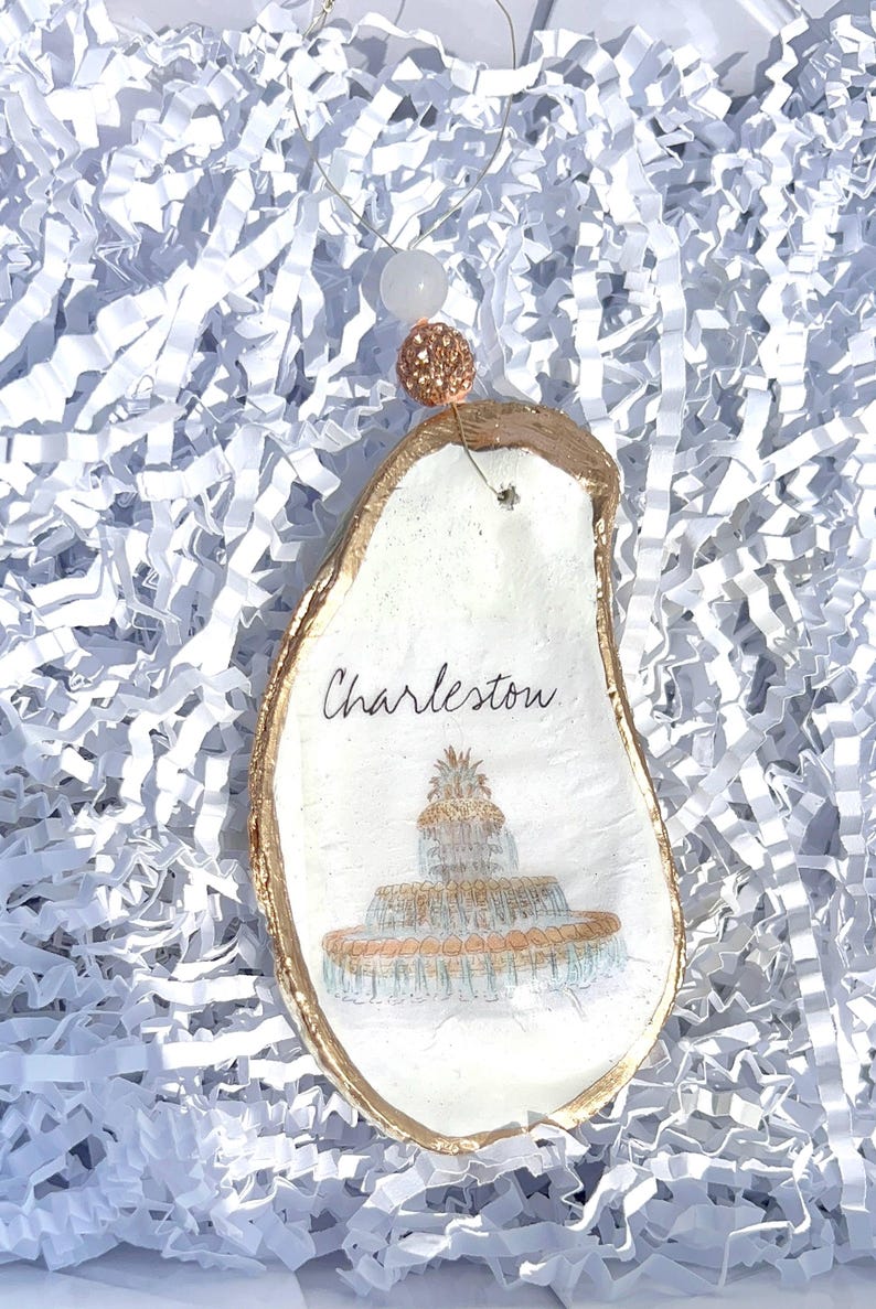 Op de afbeelding: Een witte oester schelp ornament met gouden rand, met een geschilderd beeld van de fontein in Charleston, South Carolina. De ornament is versierd met het woord "Charleston" en een gouden touwtje.