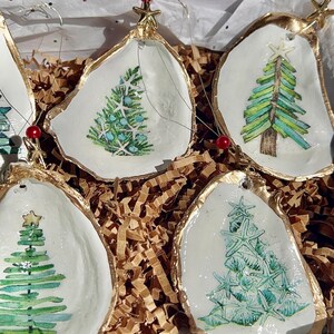 May include: Five white oyster shell ornaments with gold trim, each featuring a different hand-painted green Christmas tree design. The ornaments are nestled in brown crinkled paper.