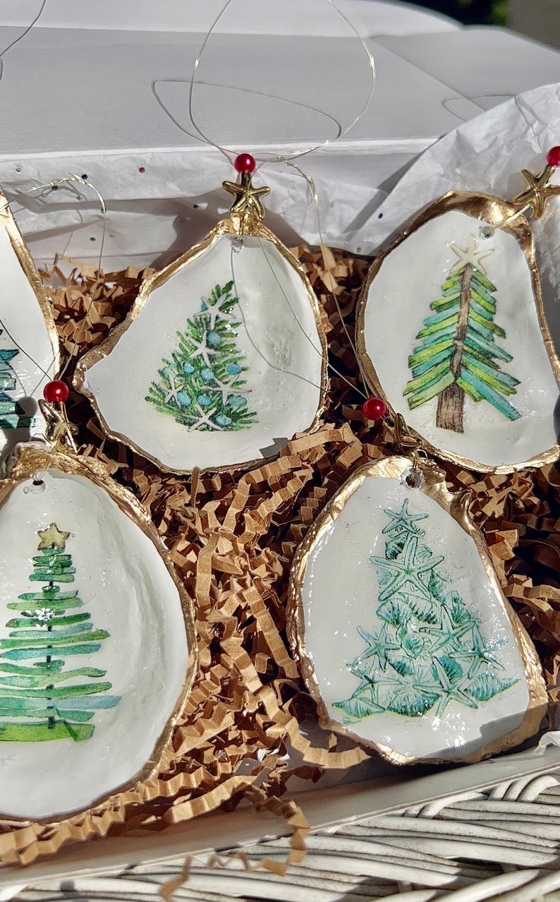 May include: Five white oyster shell ornaments with gold trim, each featuring a different hand-painted green Christmas tree design. The ornaments are nestled in brown crinkled paper.