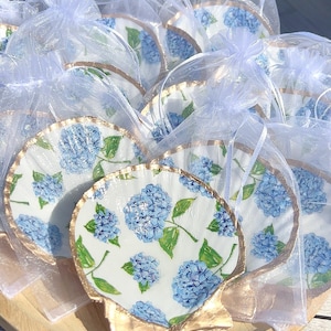 May include: A set of small, white, seashell-shaped dishes with a gold rim. Each dish is decorated with a blue and green floral pattern of hydrangeas. The dishes are displayed on a wooden surface and are packaged in individual white organza bags.