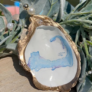 May include: A handcrafted oyster shell pendant with a gold-painted rim and a watercolor design of a blue and purple shape. The shell is adorned with a pearl and delicate wire with additional pearls, set against a backdrop of greenery.