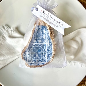 Könnte beinhalten: Eine kleine weiße Geschenkverpackung mit einer blauen und goldenen Austernschalen-Muschel im Inneren. Die Verpackung hat ein weißes Etikett mit der Aufschrift "Danke fürs Feiern der Verlobung von Kara & John 6.1.24"