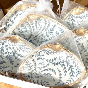 May include: A collection of decorative seashell-shaped dishes. Each dish features a white background with a blue floral pattern and gold-colored trim. The dishes are packaged in clear, sheer bags and presented in a cardboard box.