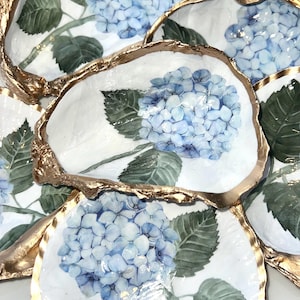 May include: A collection of oyster shells featuring hand-painted blue hydrangea flowers with green leaves on the interior. Each shell has a textured, metallic gold-painted rim, creating a decorative set of dishes with a glossy finish.