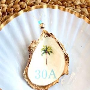 May include: An oyster shell pendant with a gold-painted edge, featuring a painted palm tree and the text "30A" in light blue. The pendant hangs from a gold cord with a blue bead and a gold bead.