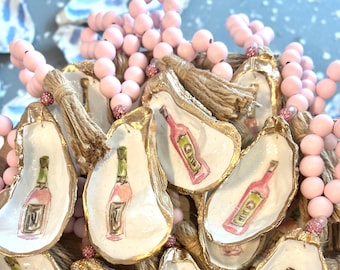 Könnte beinhalten: Eine Schnur aus rosafarbenen Holzperlen mit goldbemalten Austernmuscheln, jede mit einem handgemalten Design einer Weinflasche. Die Muscheln haben einen Quasten aus Naturbast.