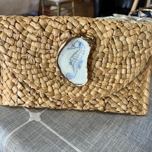 K&ouml;nnte beinhalten: Eine geflochtene Strohtasche mit einem goldenen Keramik-Seepferdchen-Design. Die Clutch ist hellbraun und hat eine geflochtene Textur.