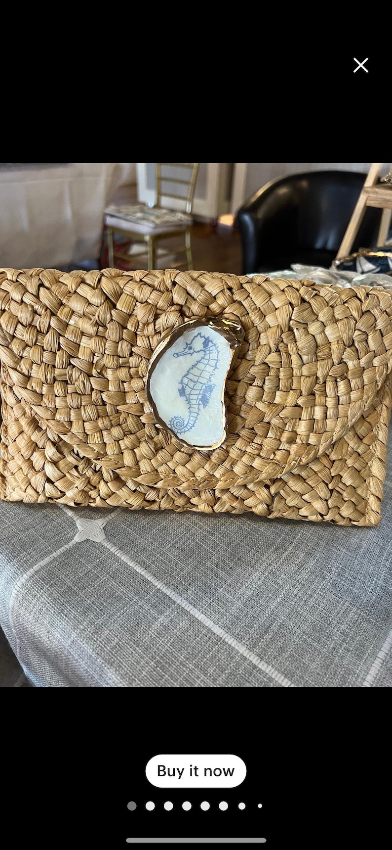 K&ouml;nnte beinhalten: Eine geflochtene Strohtasche mit einem goldenen Keramik-Seepferdchen-Design. Die Clutch ist hellbraun und hat eine geflochtene Textur.