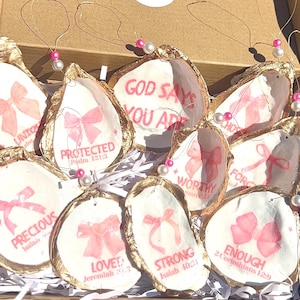 May include: A collection of oyster shell ornaments with gold edges and pink bows. Each shell features a different inspirational word or phrase in pink, such as "Protected" and "Enough", with a pearl and pink bead string.