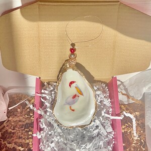 May include: A white oyster shell ornament with a gold rim and a painted pelican inside. The ornament has a red bead and gold string for hanging.