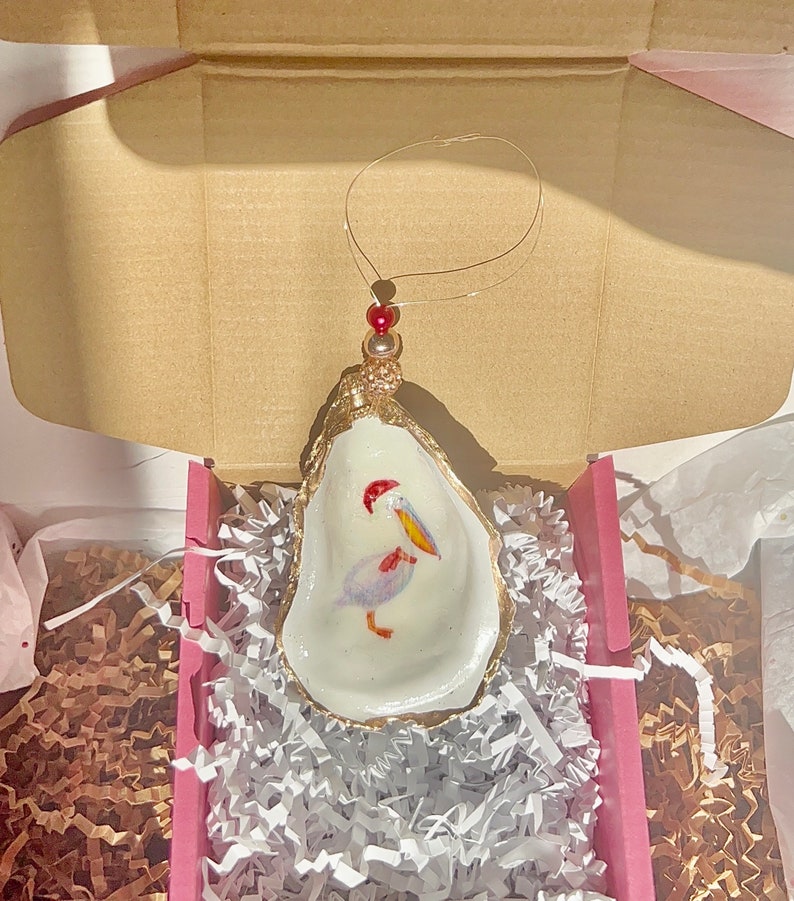 May include: A white oyster shell ornament with a gold rim and a painted pelican inside. The ornament has a red bead and gold string for hanging.