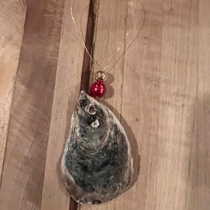 May include: A gray oyster shell ornament with a red and gold bead hanging from it. The ornament is hanging from a thin gold wire.