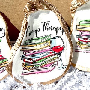 May include: A white oyster shell with gold trim, painted with a stack of colorful books, a glass of red wine, and the text "Group Therapy".