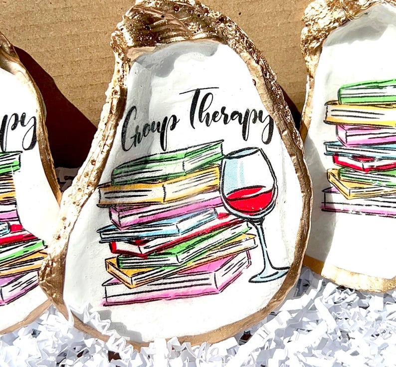 May include: A white oyster shell with gold trim, painted with a stack of colorful books, a glass of red wine, and the text "Group Therapy".