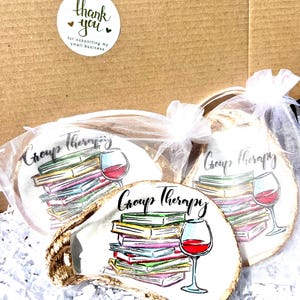 May include: Three white oyster shell coasters with a colorful illustration of a stack of books and a glass of wine. The coasters are packaged in white organza bags and feature the text "Group Therapy".