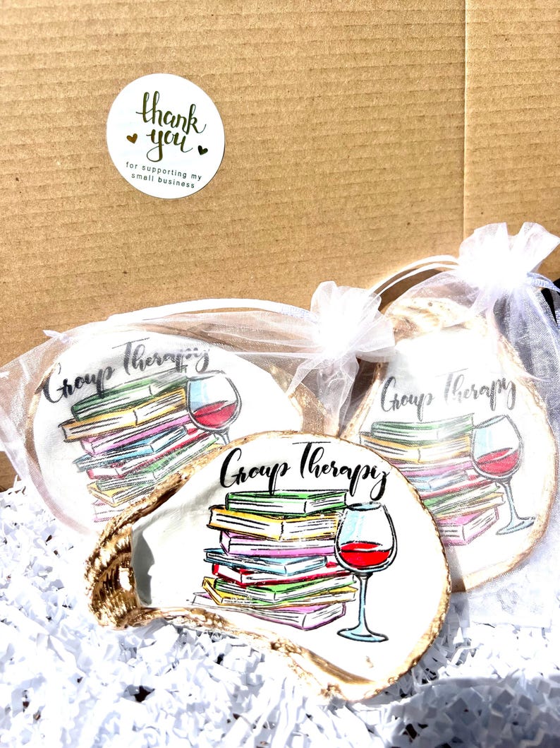 May include: Three white oyster shell coasters with a colorful illustration of a stack of books and a glass of wine. The coasters are packaged in white organza bags and feature the text "Group Therapy".