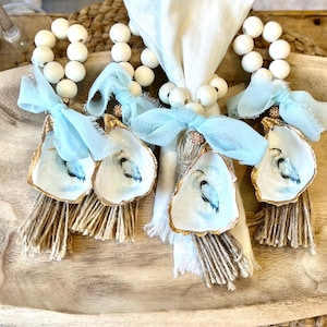 May include: Four white bead napkin rings with blue and gold oyster shell charms and blue ribbon accents. Each charm has a painted blue bird inside. The rings are displayed on a wooden tray.